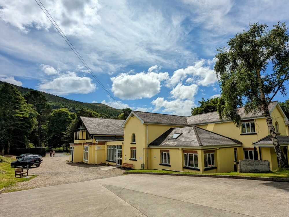 View of the exterior of a hostel in Glendalough, Ireland, with a large concrete driveway in front and a hill behind