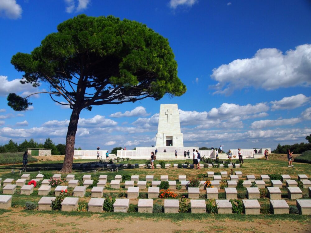 Rows of grave markers underneath a single tree, with a large memorial behind