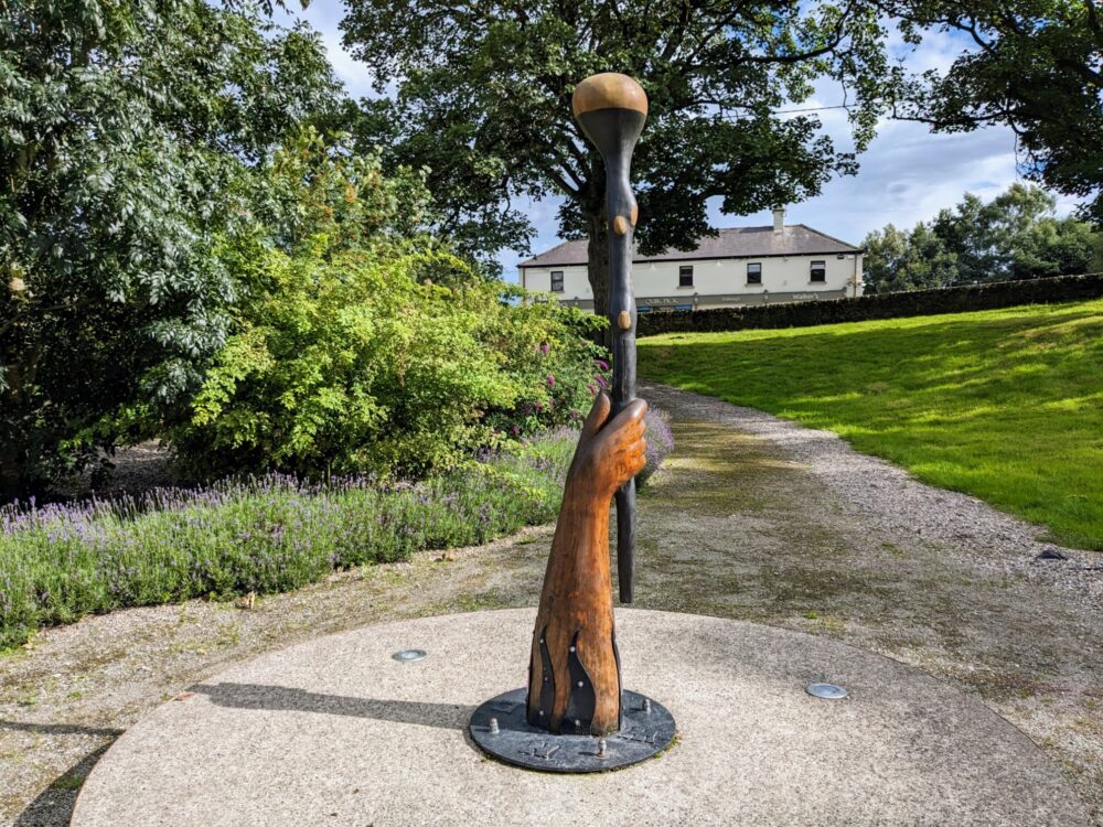Oversized sculpture in a park of an arm and hand holding a shillelagh, a traditional wooden stick used for fighting and other activities. 