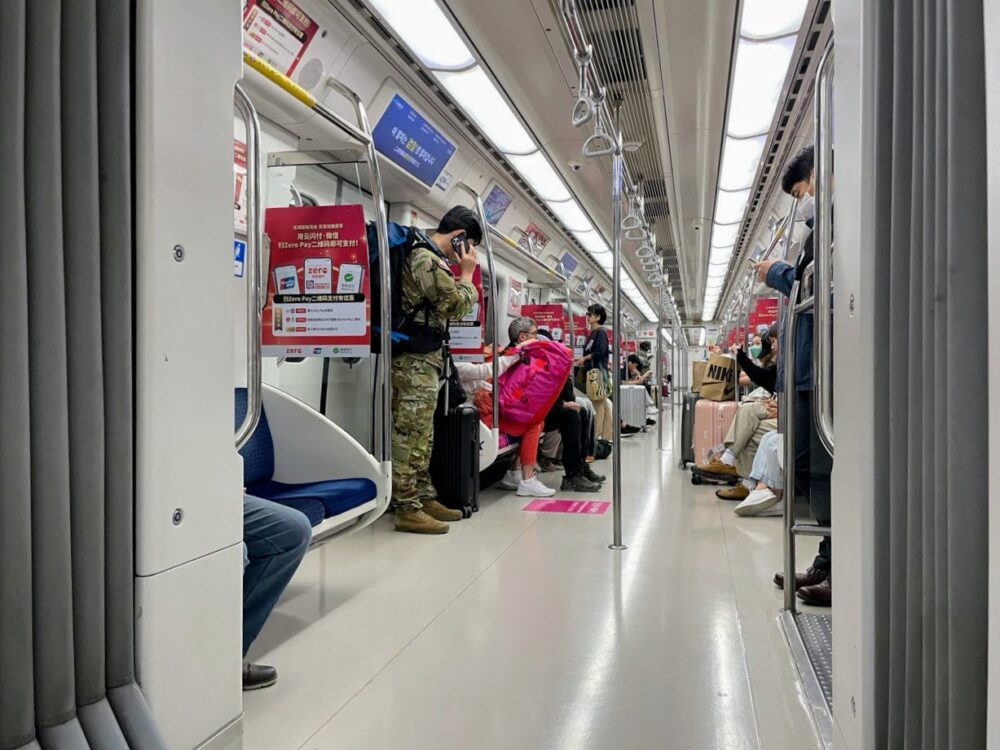 Inside of a metro train in Seoul. Several people have luggage. A man in military uniform is talking on his phone.