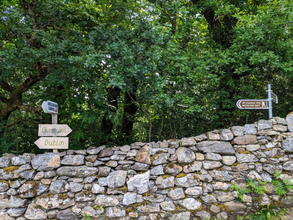 Two sets of signposts near a stone wall. One set points to Dublin, Glendalough, and Wicklow Way Camping, while the other points to the Wicklow Way in both directions.