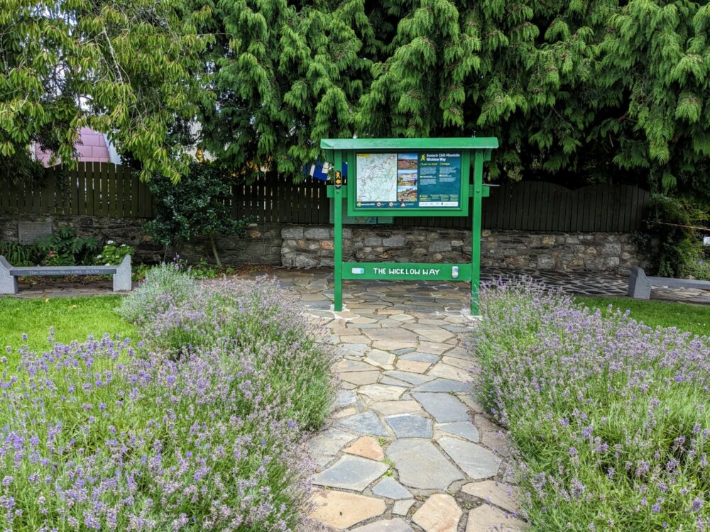Information board at the end of a short stone path with flower beds on each side. The board has "The Wicklow Way" printed on the base.