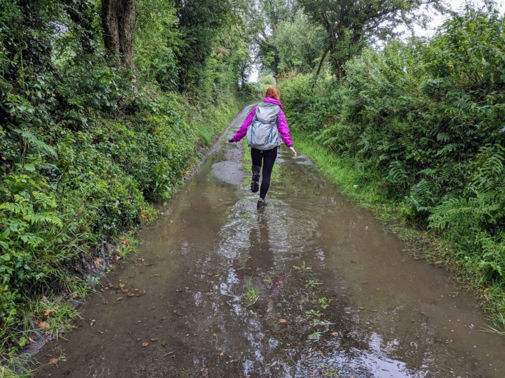 Woman wearing a backpack walking through puddles on a wet road with hedges and trees alongside
