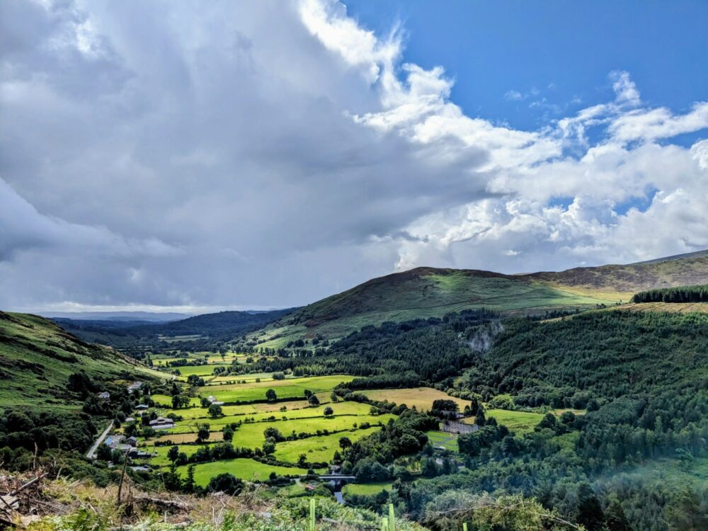 A green, sun-lit valley in Ireland, with rolling hills surrounding it. A mixture of cloud and blue sky above