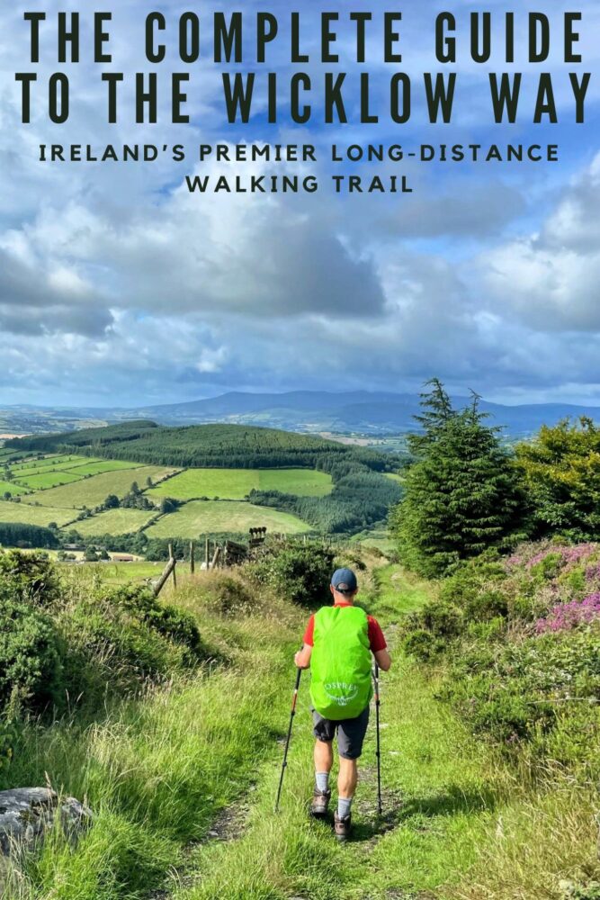 View from behind of a man with hiking poles in his hands and wearing a hiking backpack covered by a pack cover, walking along a grassy trail on a hill with expansive views over surrounding countryside, with text "The Complete Guide to the Wicklow Way" and "Ireland's Premier Long-Distance Walking Trail" overlaid at top