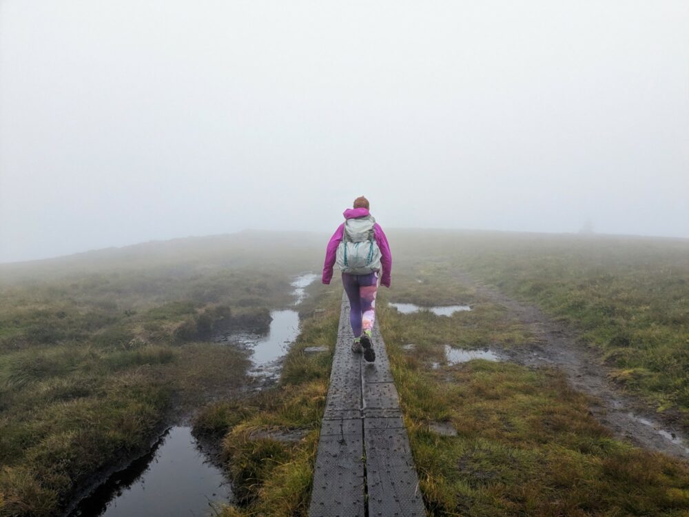 View from behind of a woman walking on a narrow boardwalk above boggy ground, with thick grey cloud all around