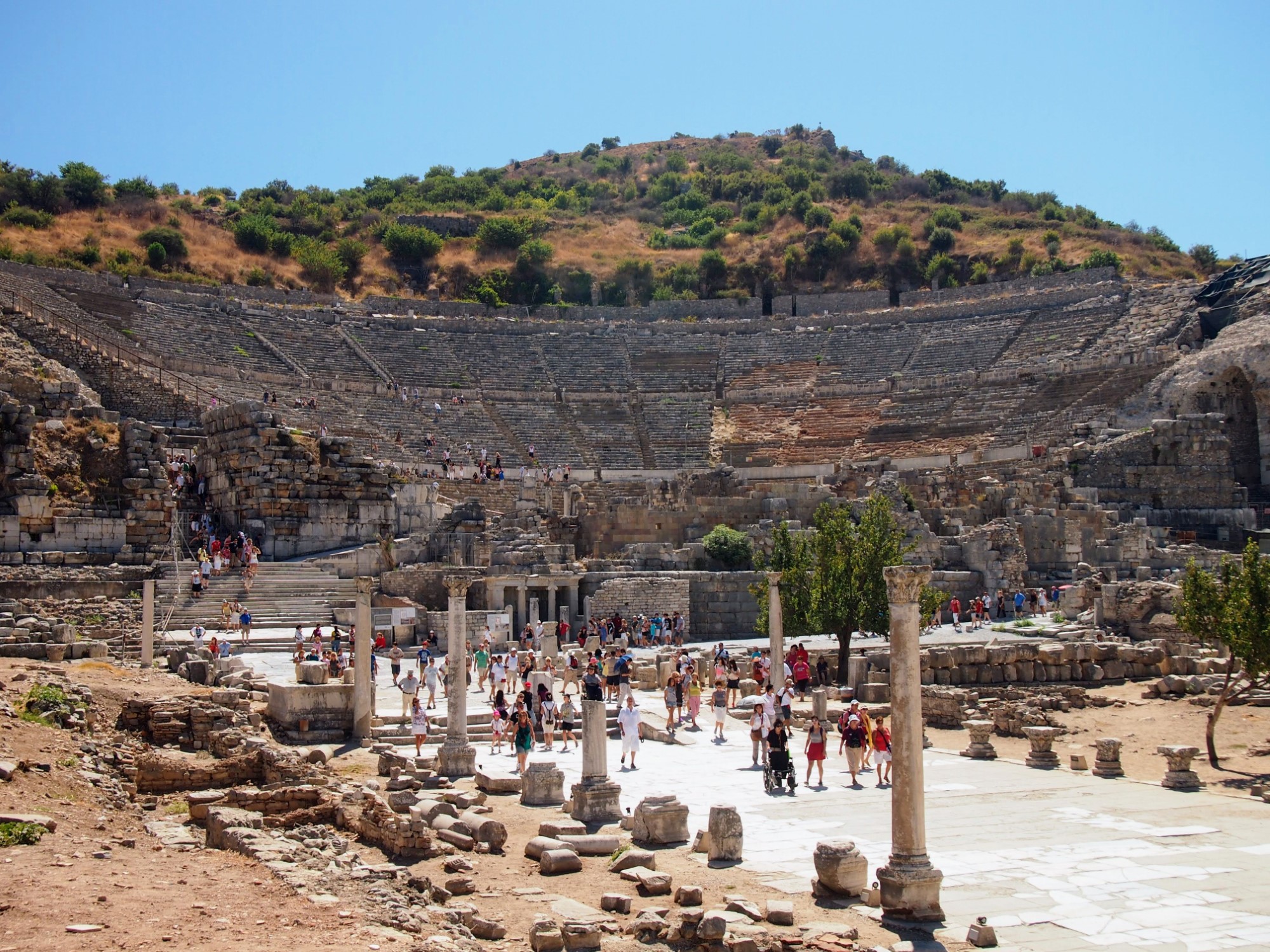 View of the ruins of a multi-storey ancient theatre at Ephesus, with people walking along a paved pathway flanked with columns in front