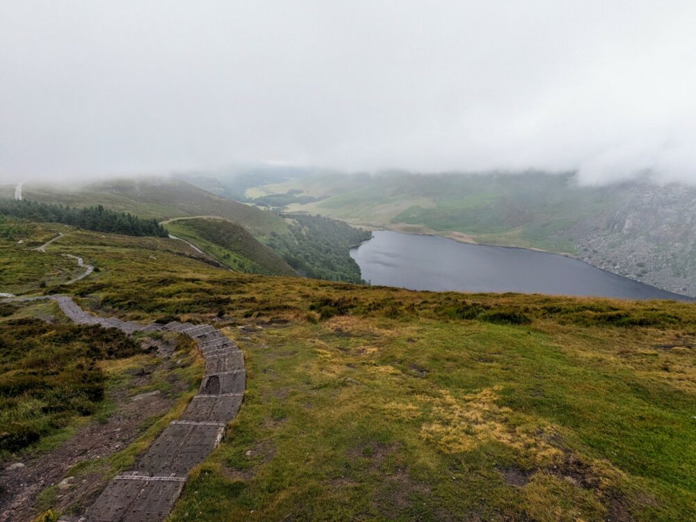 View over a lake from a high viewpoint, with thick cloud cover above. A stepped wooden boardwalk leads steeply down the hill to the left.