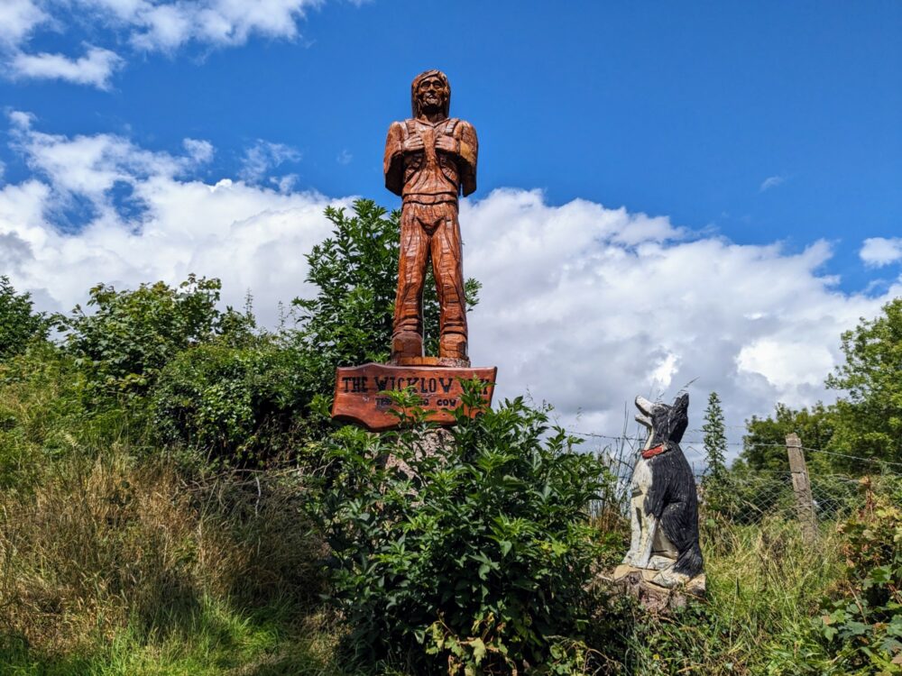 Large wooden sculptures of a man wearing a backpack with a dog alongside, and the words "The Wicklow Way" and "The Dying Cow" carved underneath