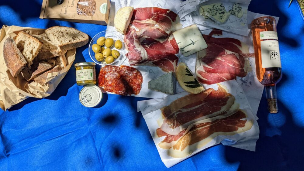 Many cured meats and cheeses laid out on paper wrappers on a picnic blanket, with sliced bread, a bottle of wine, olives, and other deli items nearby.