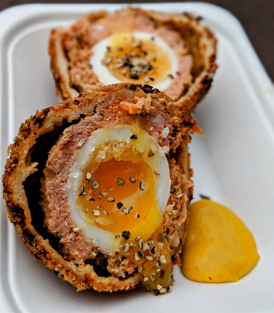 Scotch egg, cut in two with a dollop of mustard alongside, sitting on a disposable container.