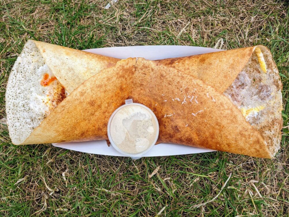 Folded dosa on a plate on the grass, with a small pot of sauce alongside