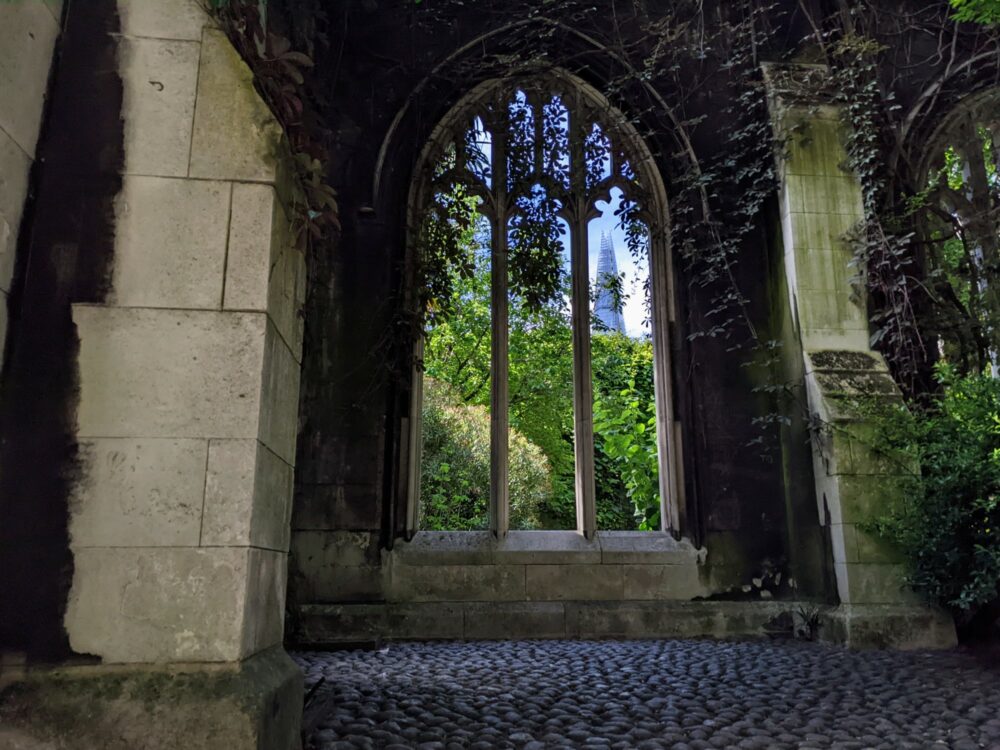 View from inside a ruined stone church with vines and trees growing up the walls, looking through a tall arched window towards The Shard in central London