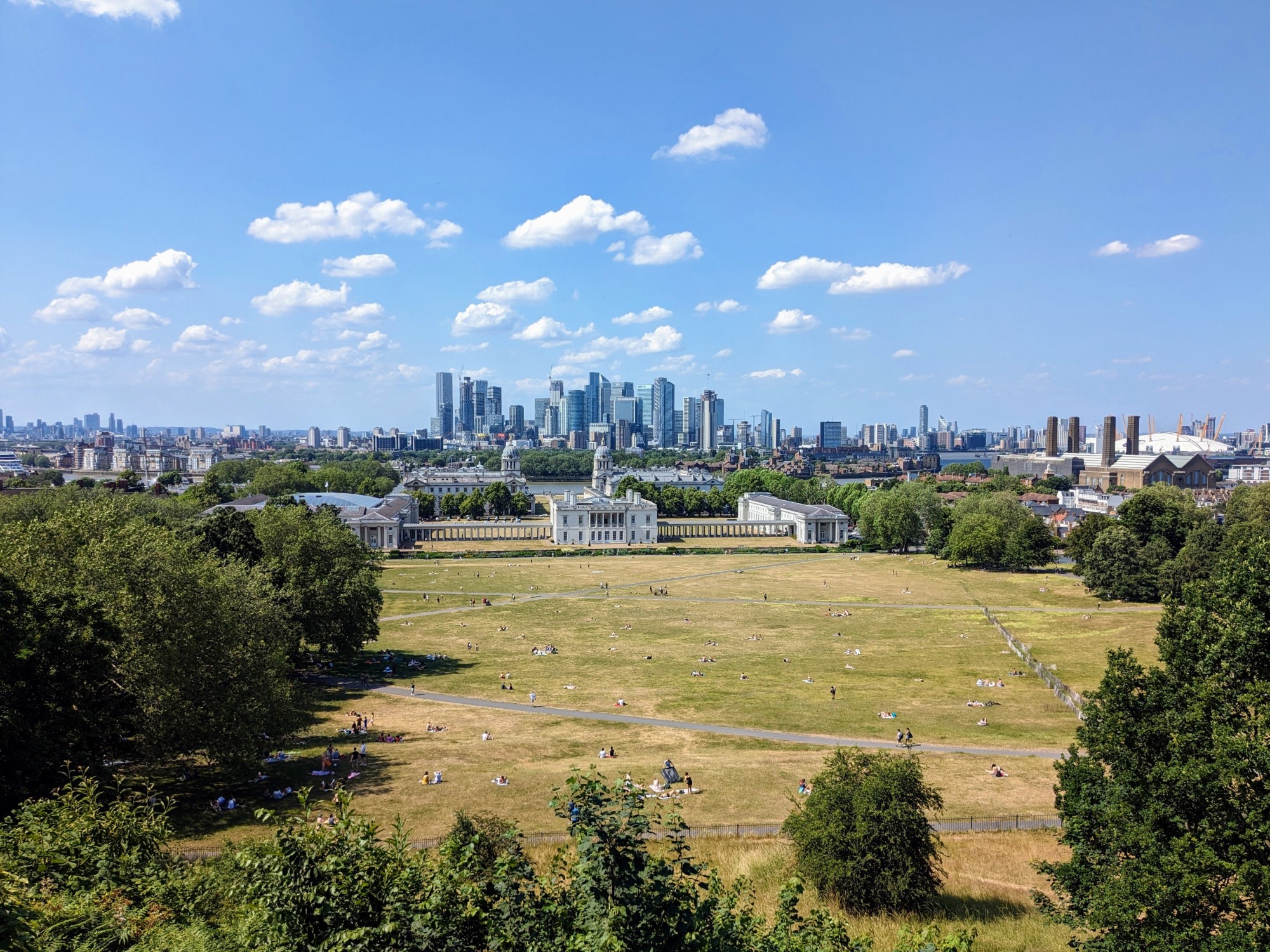 View from a hill over an expansive open grassed park with trees on either side, a grand historic building, the river Thames, and a cluster of skyscrapers beyond.