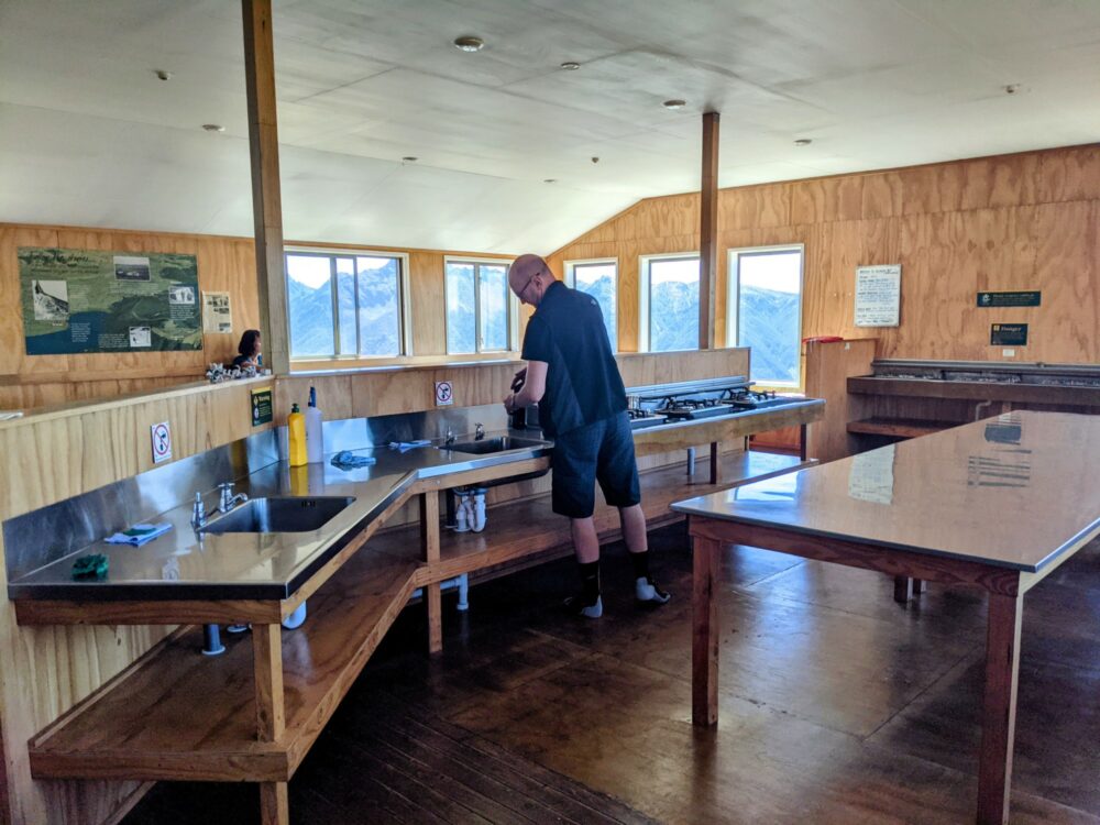 Kitchen facilities inside a camping hut (benches, sinks, gas cooktop, and a metal table), with a man stood at once of the sinks. Through the hut windows is a view of large mountains in the distance.