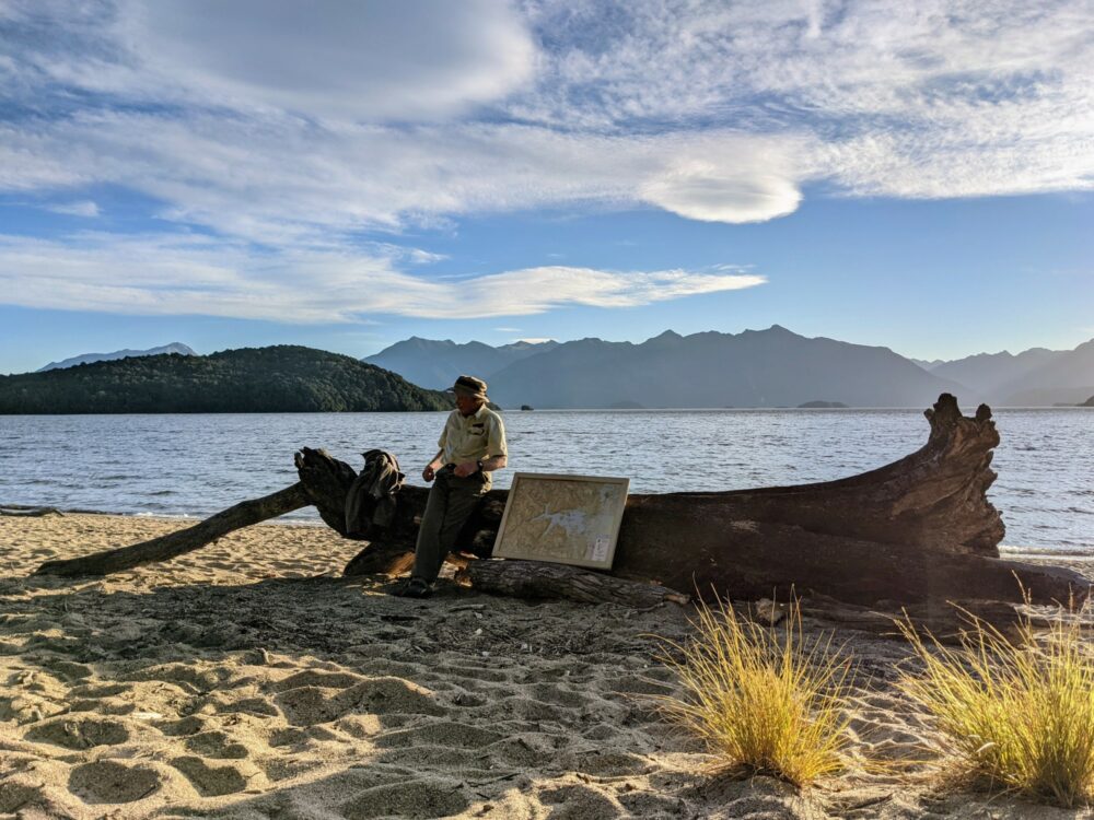 Ranger leaning up against a log on a sandy beach beside a lake, giving a talk with a large framed map propped up beside him.