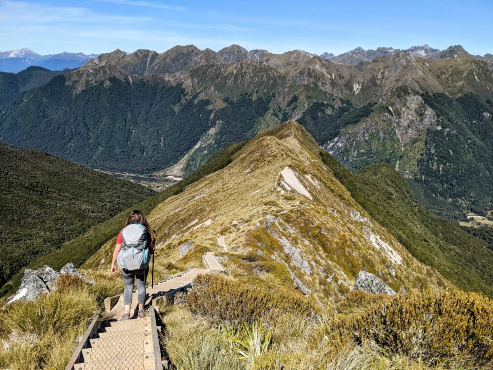 Woman walking on a long, steep wooden staircase down a mountain ridgeline, with large mountains all around.