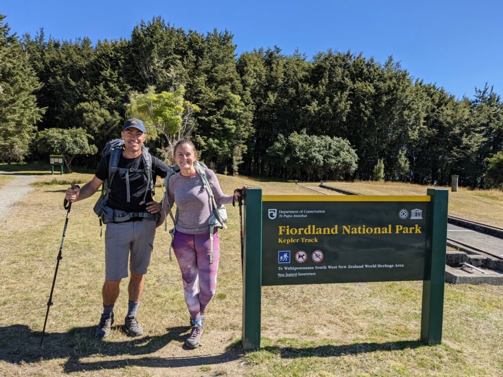 Man and woman standing beside a green and yellow sign that reads "Fiordland National Park - Kepler Track". A gravel path leads into the trees beyond.