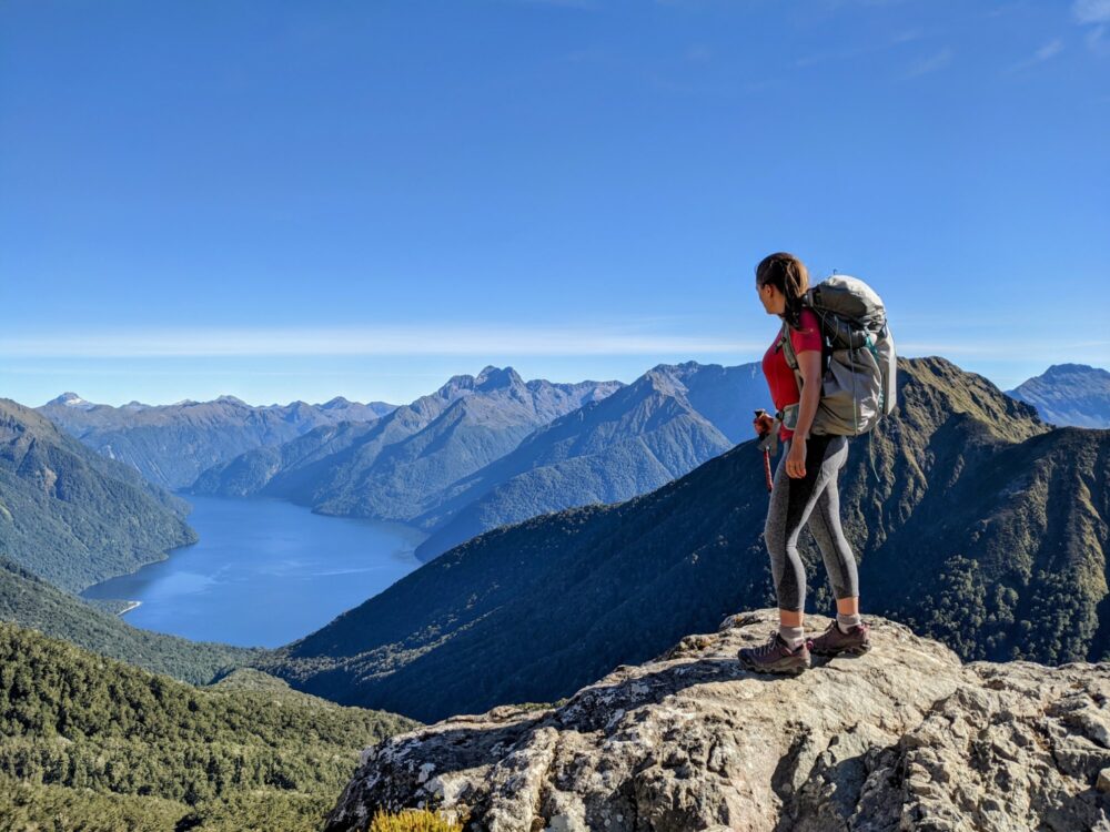 Woman wearing a backpack standing on a large rock looking down the side of a mountain towards a lake that's surrounded by mountains on all sides.
