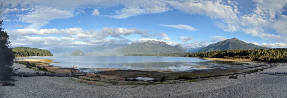 Panorama of a shallow bay in a lake with a stony beach in front and tall mountains behind