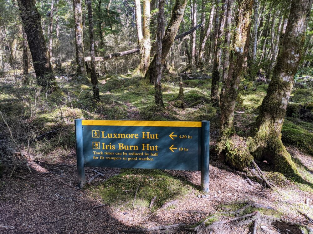 Green and yellow sign with the estimated walking times to Luxmore Hut (4.30 hr) and Iris Burn Hut (10 hr), with trees in the background.