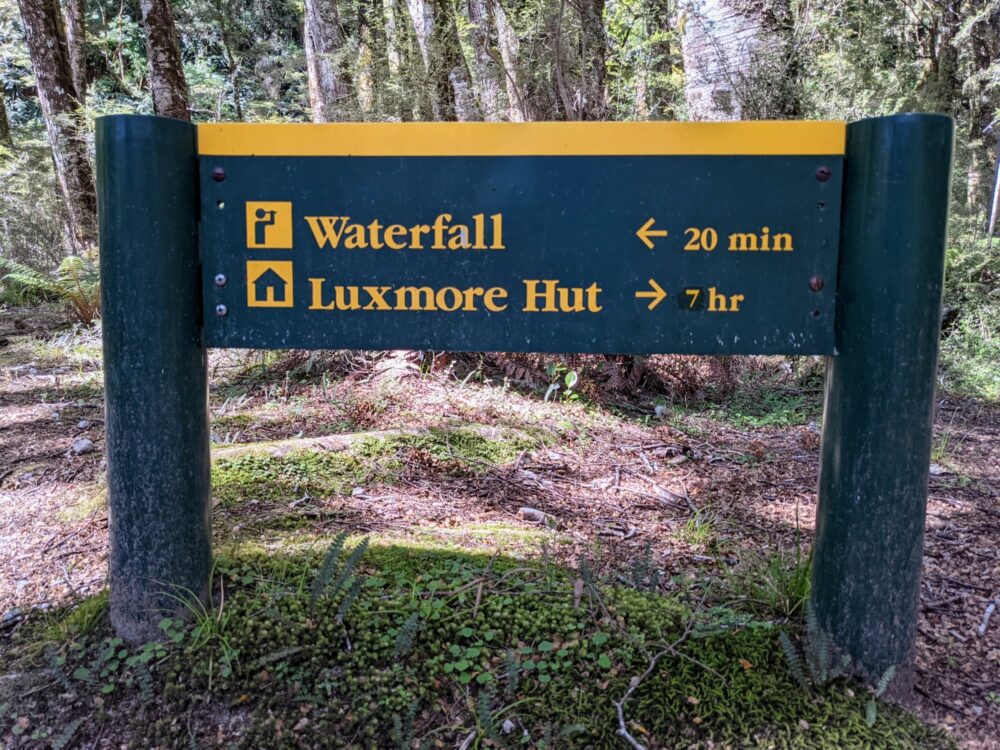 Green and yellow sign showing walking times to a waterfall (20 minutes) and Luxmore Hut (7 hrs), with trees behind