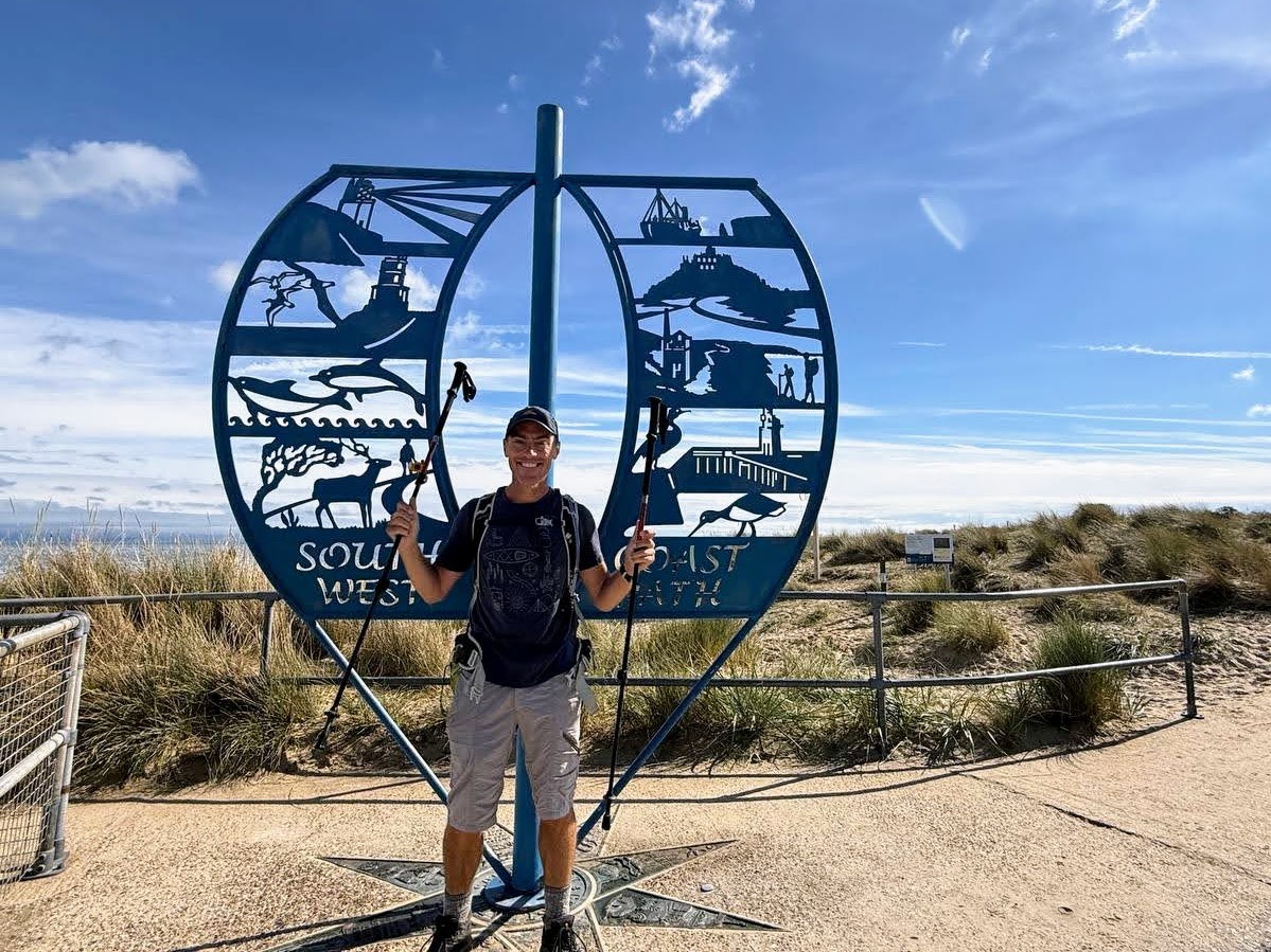 Man in hiking clothes, holding walking poles and wearing a backpack, standing in front of a blue metal sculpture with stylised images and the words "South West Coast Path"
